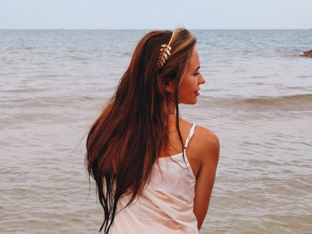 KaraCriegs, con lunghi capelli castani, indossa una canotta bianca e bagnata, senza maniche, ed è in piedi in acque poco profonde dell'oceano, guardando di lato, tenendo in mano una parrucca dorata..