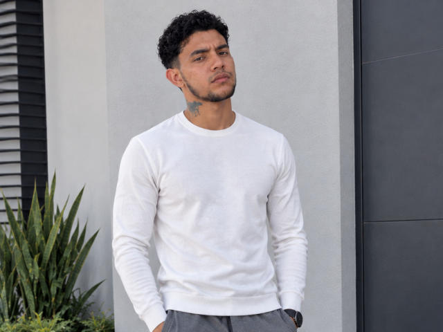 MasterEmiliano with curly black hair, white long-sleeve shirt, gray pants, hands in pockets, tattoo on left neck, standing, serious expression, light skin, green eyes, and slight beard.