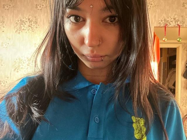 TrenaShad mit langen, dunklen Haaren, Nasenring und Lippenpiercing, trägt ein blaues Hemd mit einem gelben Aufdruck und blickt in die Kamera. Braune Augen, hellbraune Haut..