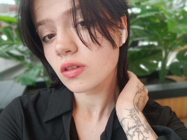 Shanika Castanon, con cabello negro corto, labios rosados, audífonos blancos, camisa negra, tatuaje en la mano derecha, mirando ligeramente hacia un lado, con la mano cerca del cuello..