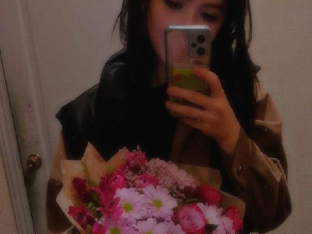 EleanoraMirabal with dark hair, holding a phone, wearing a brown jacket, taking a mirror selfie, showing a bouquet of pink and white flowers, with a black scarf.