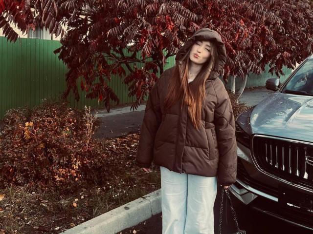 EleanoraMirabal with long brown hair, wearing a brown puffy jacket, gray cap, and light blue pants, standing next to a black BMW, red-leaved tree in the background.
