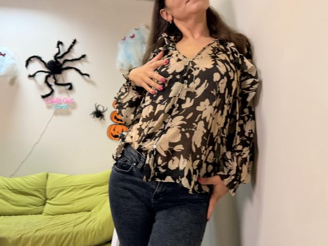 Tilliana with long brown hair, wearing a beige floral blouse and dark jeans, touching her chest, standing against a wall, with pink nails and a confident pose.