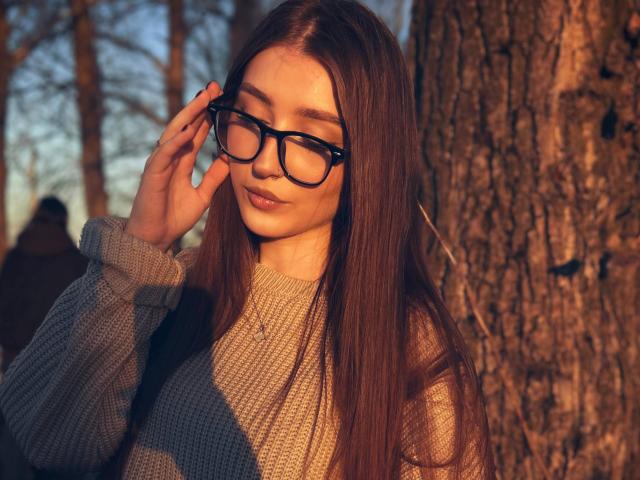 Eleanora Mirabal, con lunghi capelli castani, che indossa occhiali neri e un maglione grigio, tocca gli occhiali con la mano destra, mentre la luce del sole proietta ombre calde sul suo viso..