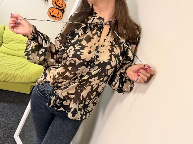 Tilliana, com uma blusa floral marrom, abotoada até a gola, calças escuras, segurando um barbante, unhas cor-de-rosa, com a cabeça fora do quadro, em um quarto com uma cadeira verde e decorações de Halloween..