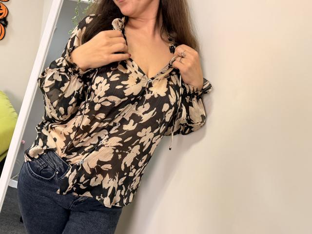 Tilliana, com longos cabelos castanhos, vestindo uma blusa preta com estampas florais, ligeiramente aberta para mostrar seus pequenos seios, e jeans azuis, apoiada em uma parede branca, segurando a blusa..