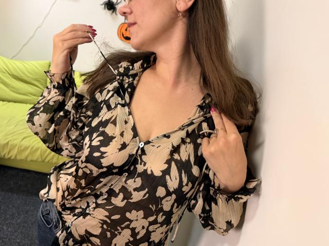 Tilliana, com cabelo castanho, vestindo uma blusa floral transparente, ligeiramente desabotoada, mostrando pequenos seios, segurando um pirulito de Halloween, com unhas cor-de-rosa, apoiada em uma parede branca..