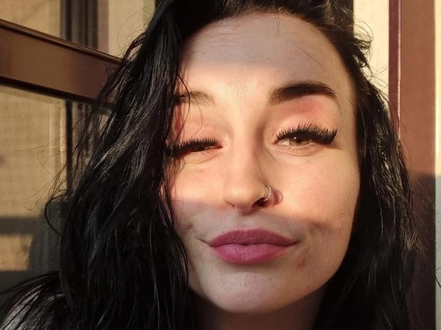 LucySensitive a les cheveux noirs ondulés, un anneau de nez et du rouge à lèvres rose, elle cligne des yeux avec de longs cils et affiche un léger sourire, créant une ombre sur sa peau pâle..