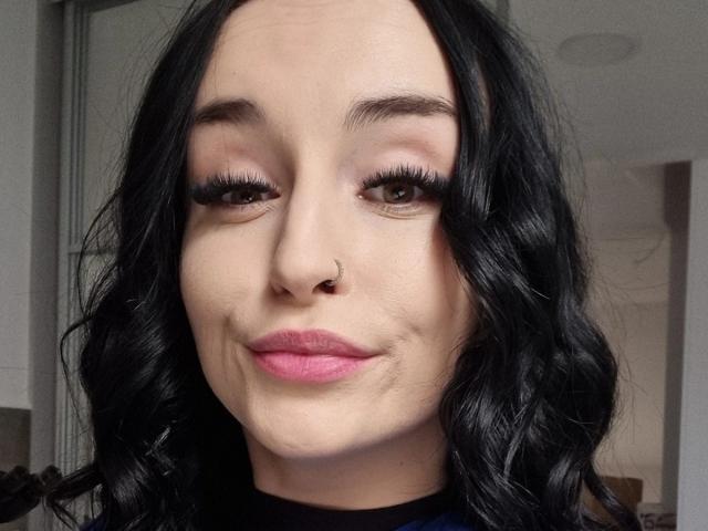 LucySensitive tem cabelo preto ondulado, olhos castanhos, cílios longos, batom rosa e um pequeno piercing no nariz. Ela está sorrindo levemente, com um rosto bonito e atraente..