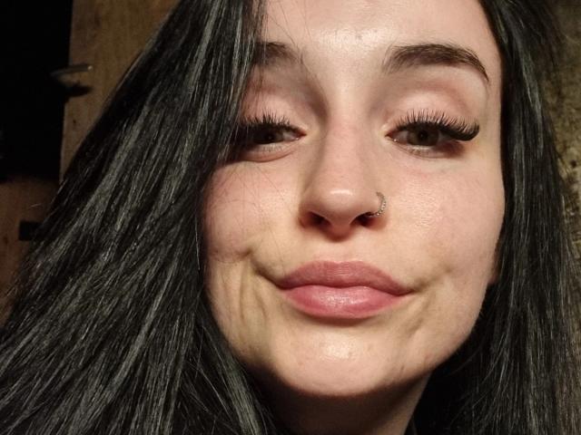 LucySensitive tiene la piel clara, cabello lacio y oscuro, ojos marrones con pestañas largas, un piercing en la nariz y labios rosados con finas líneas de expresión alrededor de la boca. Su rostro está centrado en la imagen..