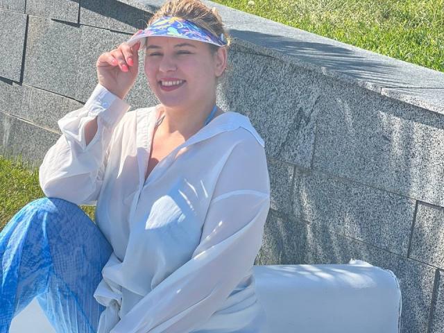AvaQueen, with blue eyes and blonde hair, wears a sheer white blouse and blue pants, smiling, holding a colorful headscarf, sitting against a grey stone wall.