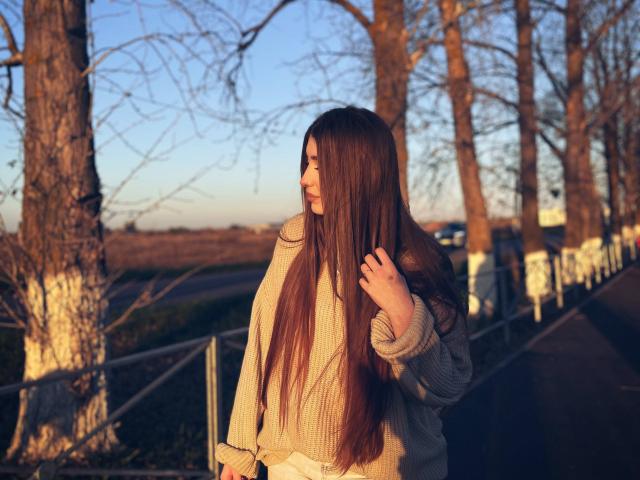 EleanoraMirabal with long brown hair, wearing a beige sweater, standing on a sunlit path, hand in hair, looking to the side, golden hour, trees in background.
