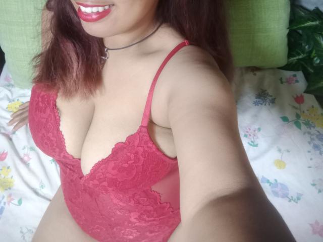 MariahSquirting69 con lencería roja de encaje, mostrando sus grandes senos, sonriendo con lápiz labial rojo, collar y cabello rojo, tomando un selfie, acostada en una cama con estampado floral..