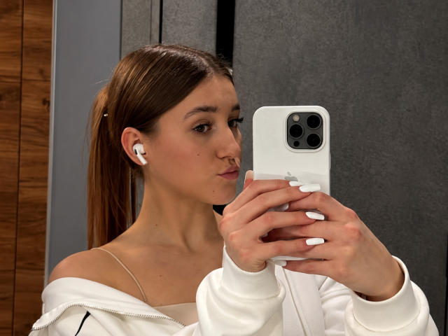 TeneshaCisco con una coleta castaña, sudadera blanca con un hombro descubierto, auriculares blancos, tomando un selfie con un iPhone blanco, expresión de labios fruncidos, uñas blancas, una pared de madera parcialmente visible..