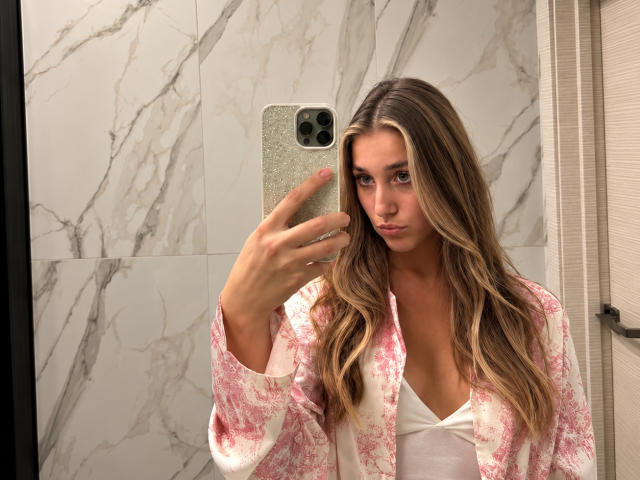 TeneshaCisco with long brown hair, pink and white floral robe, white V-neck, holding glittery phone, taking mirror selfie, showing subtle makeup, looking serious, in bathroom.