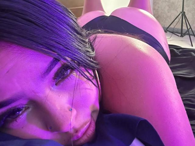 LiliMilk lying on her stomach, black thong, purple lighting, visible butt, long dark hair, silver nose ring, serious expression, close-up, focus on face and upper body.