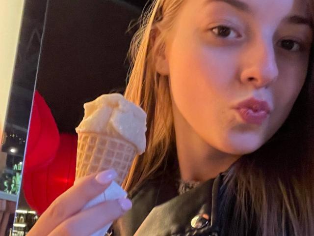 DorlaSinibaldi with pouty lips, holding a vanilla ice cream cone in her right hand, wearing a denim jacket. She has long brown hair and light skin.