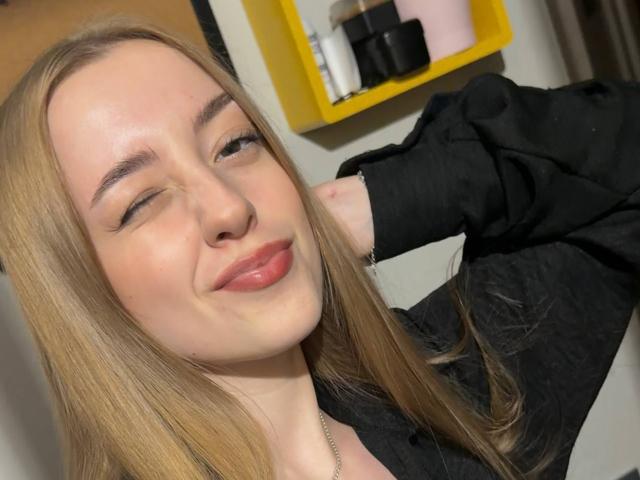 DorlaSinibaldi with blonde hair, black shirt, and pink lips, winking and smiling with her hand behind her head, showing off her cute face and small ass.