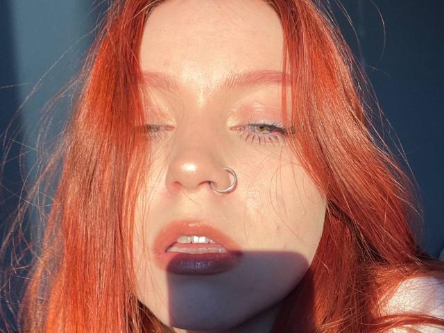 LilliamChevalier with red hair, nose ring, shiny pink lips, and closed eyes in sunlight, showing her shiny nose and soft freckles, with a slight shadow on her face.