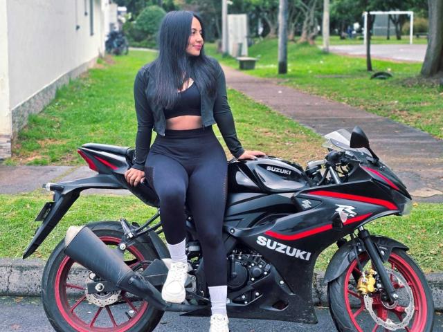 IsaLawrence with long black hair, black sports bra, high-waisted black leggings, white socks, and white sneakers, sitting on a black Suzuki motorcycle with red accents.