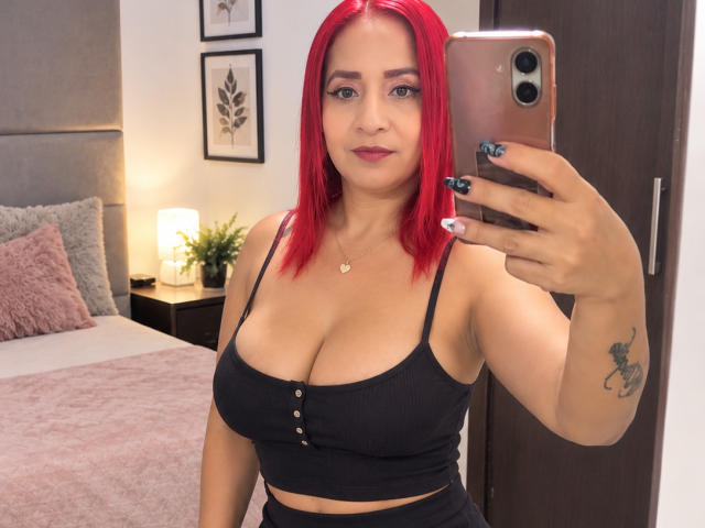 KateMonroe, avec ses cheveux roux, un haut noir qui met en valeur sa poitrine généreuse, tenant un téléphone, prenant un selfie devant un miroir, a un tatouage sur le bras et porte un petit collier..