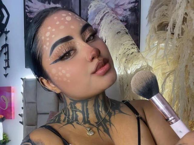 LairaHearvy, com tatuagens, maquiagem facial clara com manchas, sobrancelhas pretas, batom rosa, segurando um pincel rosa, blusa preta com alças, colar e cabelo escuro preso, olhando para a câmera..