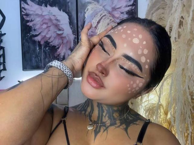 LairaHearvy mit weißem Punkt-Make-up im Gesicht, geschlossenen Augen und einem Lächeln, die sich mit der Hand am Kopf berührt, wobei ein tätowiertes Handgelenk und ein silbernes Armband zu sehen sind, trägt ein schwarzes Oberteil..