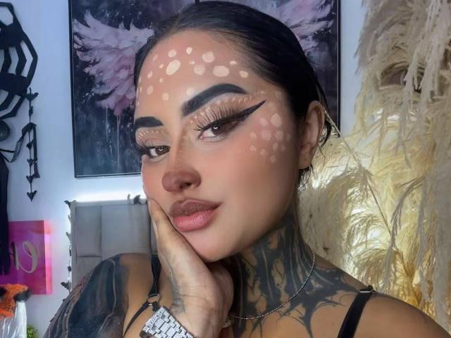 LairaHearvy tem cabelo escuro, tatuagens e manchas brancas em formato de coração no rosto. Ela está tocando o queixo, com um delineador dramático e lábios rosados. A mão dela está na bochecha..