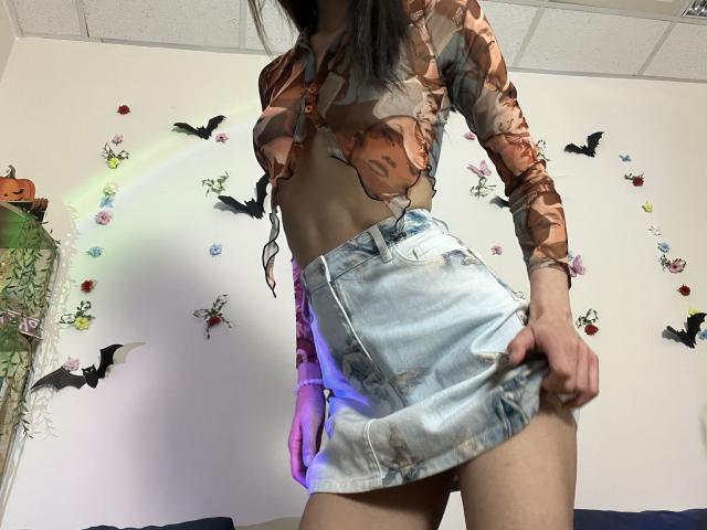 Valnes with long black hair, brown floral top showing tits, and blue denim skirt, pulling up skirt to show bare ass, standing in a room with Halloween decor.