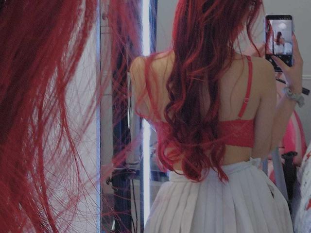 CherryBuchel with long red hair, red bra, white pleated skirt, taking a selfie, showing her back, holding a phone, mirror reflection, red bra straps, wavy hair.