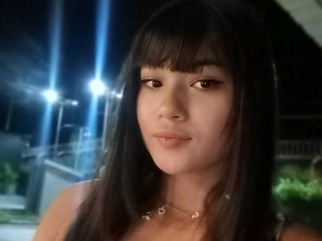 AshlyLee has straight black hair with bangs, brown eyes, and a subtle smile. She's wearing a heart necklace, and her cute face is centered in the dimly lit photo.