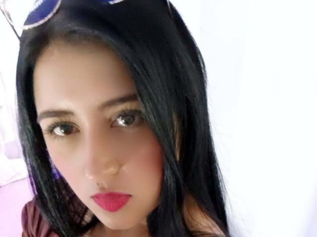 NicollJeans has black hair, brown eyes, and red lipstick. She's wearing sunglasses on her head and has a small nose ring. Her face is slightly blurred.