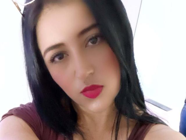 NicollJeans has dark black hair, red lipstick, and a slightly flushed face, looking at the camera with a neutral expression. She's wearing a maroon top.