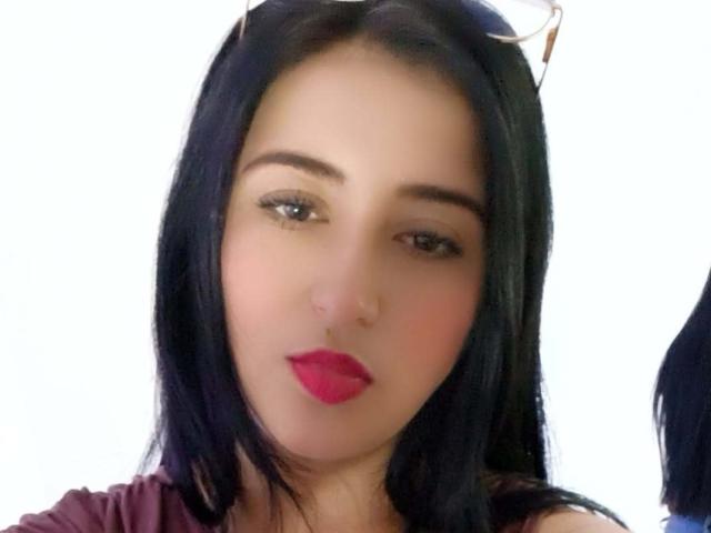 NicollJeans has dark, straight hair, light skin, red lipstick, and brown eyes. Eyeglasses rest on her head. She's wearing a pinkish shirt. Her ass is not visible.