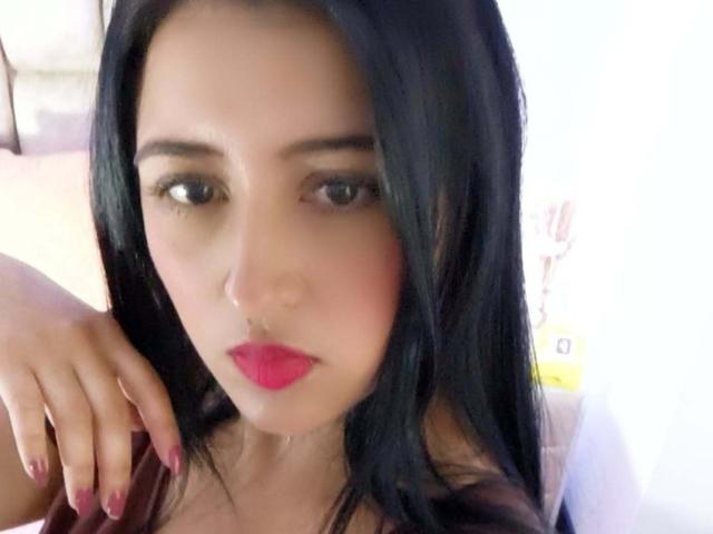NicollJeans has black hair, pink lipstick, and dark eye makeup, with a hand resting near her face. Her nails are painted red. She has a serious expression.