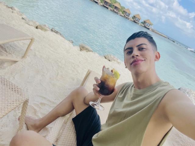 ImMateo, met kort donker haar, een groen mouwloos shirt en zwarte korte broek, zit op een strandstoel, met een drankje met ijs in zijn hand, en glimlacht naar de camera..