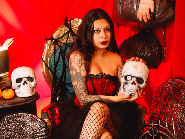 GabyJoy with tattoos, wearing a red and black corset, fishnet stockings, holding a skull, sitting on a throne, with another skull and Halloween decor. Boobs, ass, tattoos.