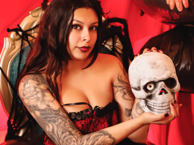 GabyJoy with dark hair, tattoos on her right arm, holding a skull, wearing red and black lingerie, looking at the camera with a slight smile.