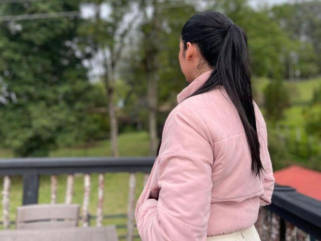 MilusSky with black ponytail, wearing pink fuzzy jacket, standing with arms crossed, facing away, showing ass in white pants, on wooden balcony.