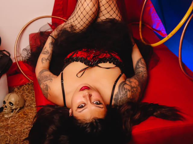 GabyJoy lying upside down on a red couch, wearing black fishnet stockings, red and black lace lingerie, and a black tutu, showing her tattoos and red lipstick.
