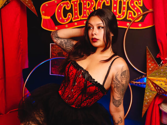 GabyJoy with tattoos, red-black corset, black tutu, and red lipstick, sitting with arm behind head, looking at camera, showing her tits and ass. Circus-themed setting.