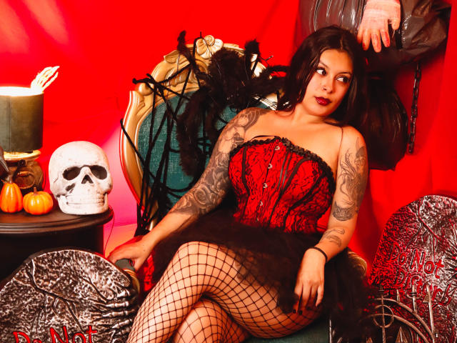GabyJoy with tattoos, red-black corset, black fishnet, and black skirt, sitting on ornate chair, showing ass, with skull, pumpkin, and lace hand on red background.