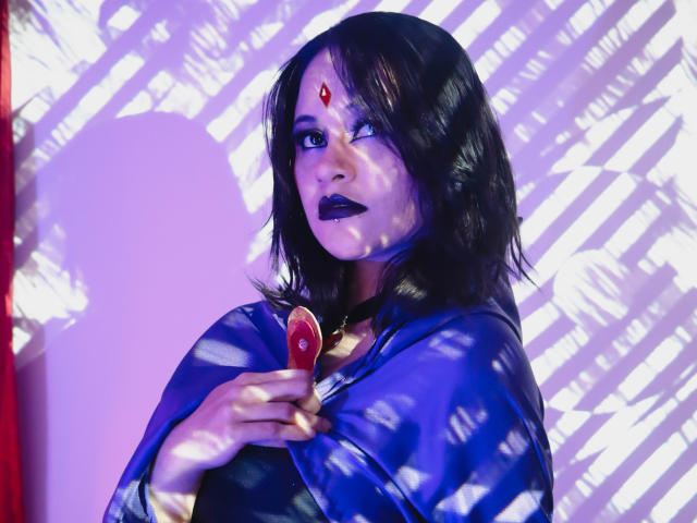 CassieJones with black hair, purple lipstick, and a red gem on her forehead, holding a red object, wearing a blue garment, shadowed by diagonal white lines.