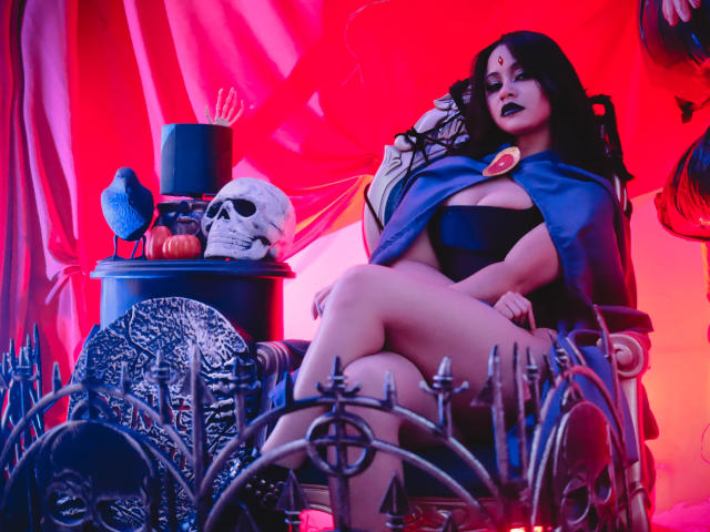 CassieJones with black lipstick, dark hair, blue cape, black bra, and black panties, sitting on a throne, legs crossed, skull on a table, red and pink lighting.