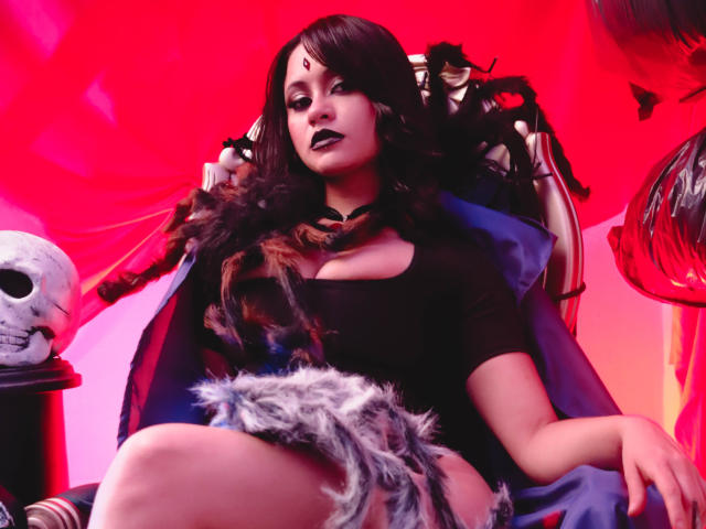 CassieJones with dark hair, black top, and furry shorts, sitting on a throne with a skull, red background, and black lipstick, looking confident. Boobs visible under top.