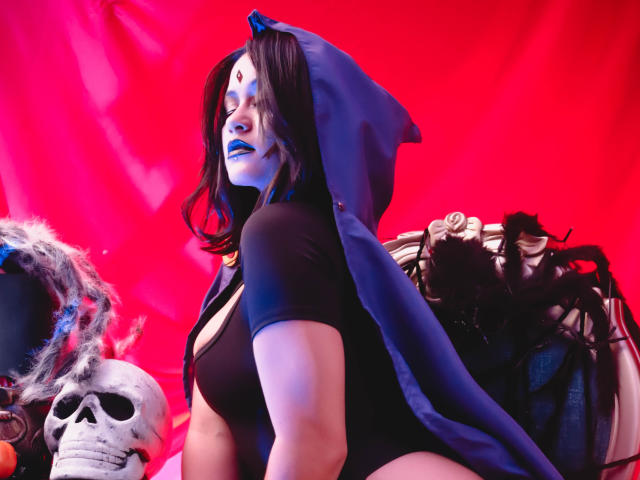 CassieJones in a black bodysuit, blue hooded cape, white face paint, blue lipstick, and a red forehead dot. Skull on left, black mask with horns on right.