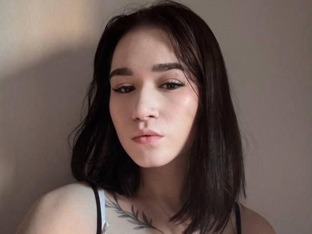 MaryanWelshans has pale skin, black shoulder-length hair, and wears a black spaghetti strap top, showing a tattoo on her chest. She has thick eyebrows and subtle makeup.