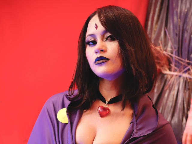 CassieJones with purple makeup, black choker, red heart pendant, and purple shirt, showing her tits, has dark brown hair, and a red jewel on her forehead.