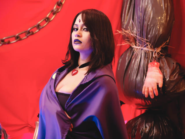 CassieJones with dark brown hair, purple cloak, black top, blue lipstick, red gem necklace, and third eye mark, standing next to a bagged, bound arm.