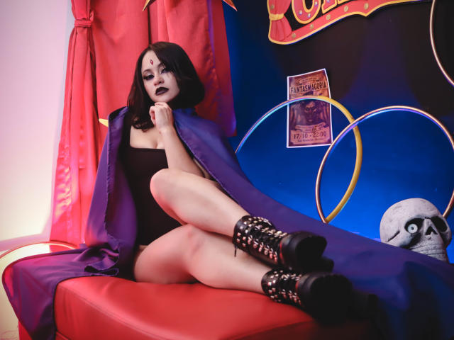 CassieJones with black bob, dark lipstick, black strapless top, purple cape, black boots with studs, sitting on red bench, legs crossed, showing pale skin and black panties.